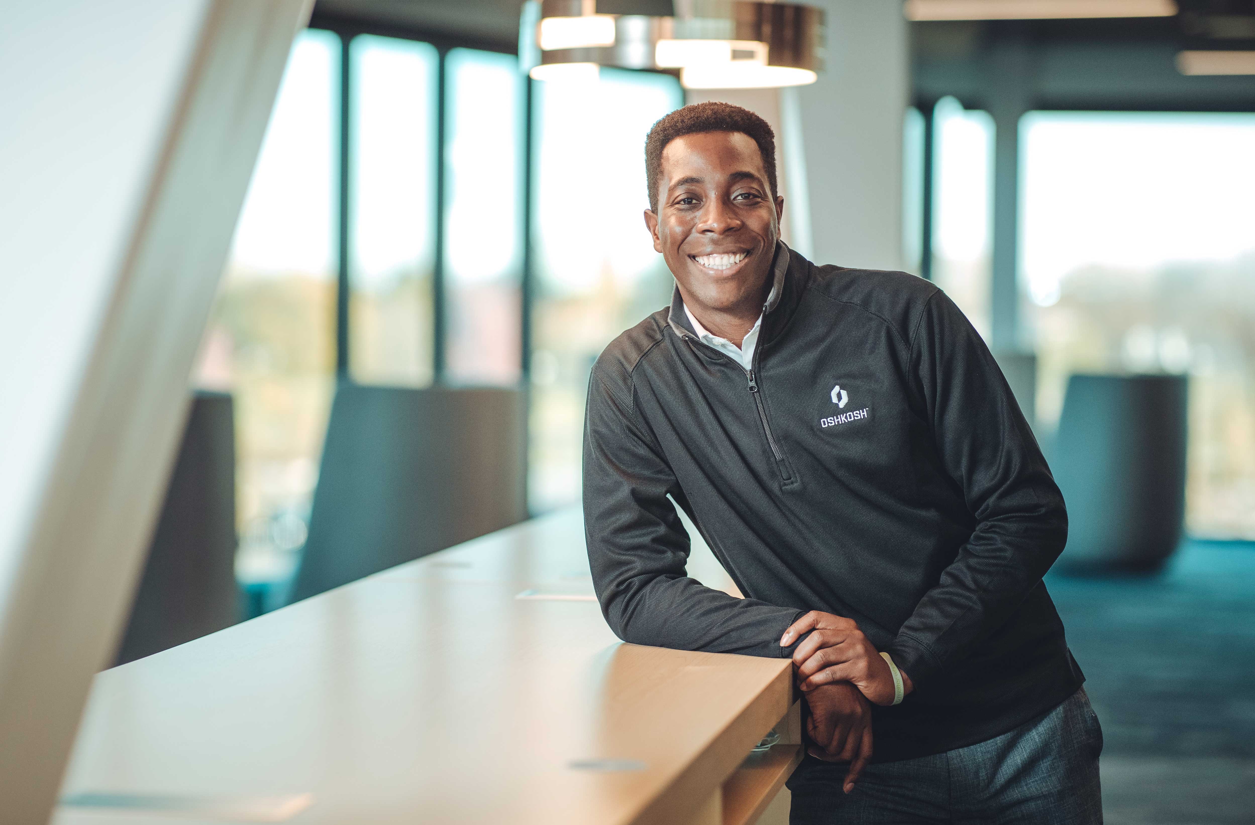 Young black man wearing a black Oshkosh 1/4 zip sweatshirt standing at a tall table in an office setting